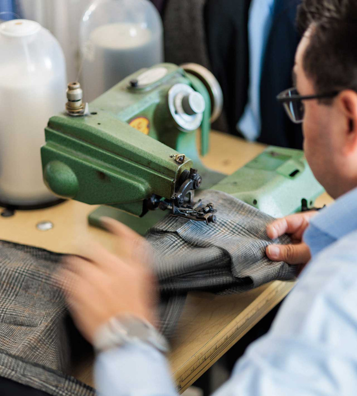 A close-up lifestyle shot of a man in a light blue shirt and glasses operating a green industrial sewing machine to sew gray plaid fabric.