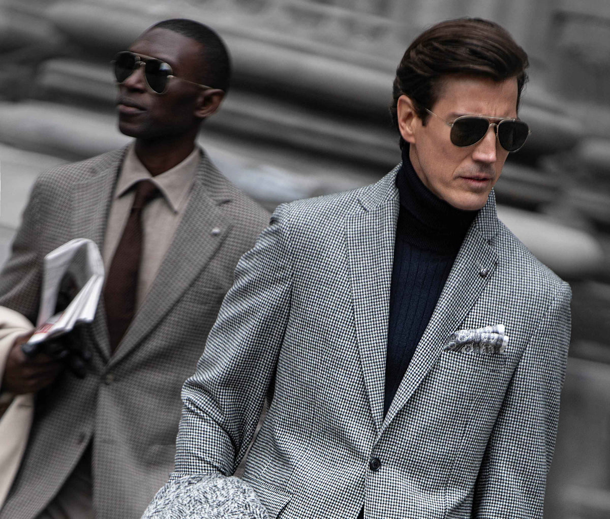 Two men in tailored suits wearing aviator sunglasses, photographed outdoors against a blurred stone or concrete background. The foreground man wears a light gray and white micro-check suit with a dark navy turtleneck and a patterned pocket square, while the second man behind him wears a taupe suit with a brown tie.