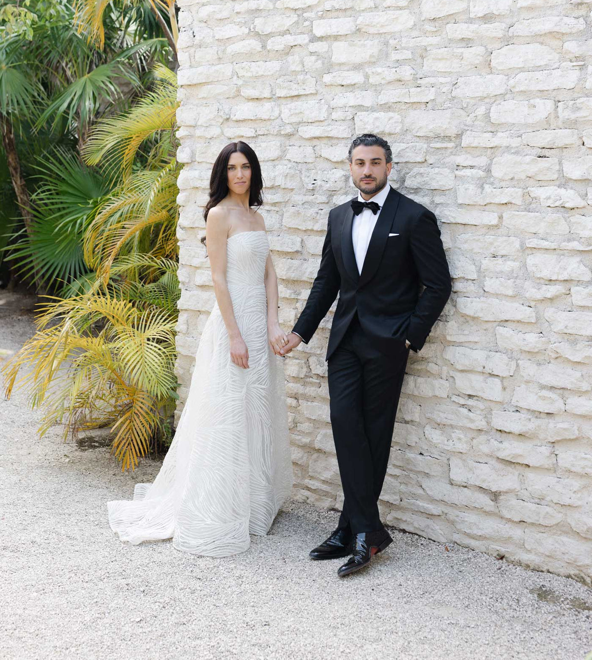 A bride in a strapless white wedding gown and a groom in a black tuxedo holding hands, standing outdoors against a light stone wall with palm foliage on the left.
