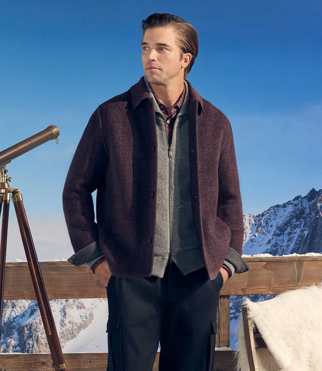 A man stands outdoors in a snowy mountain setting wearing a dark brown textured jacket over a gray zip sweater, plaid shirt, and dark pants. A wooden fence, telescope, and chair are visible in the scene.